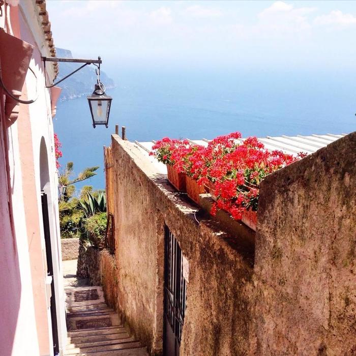 Narrow stone stairway in Ravello with stucco walls, red flowers, and the blue sea beyond