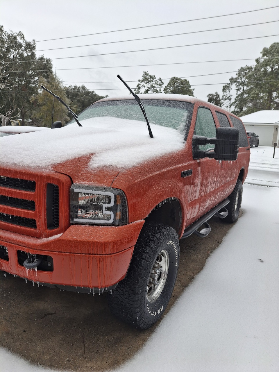 Red Ford pickup truck with snow on hood and roof parked in snowy driveway with trees behind
