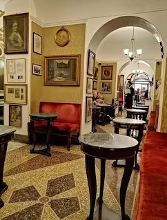 Antique cafe interior with red benches, marble tables, framed paintings, and arched hallways