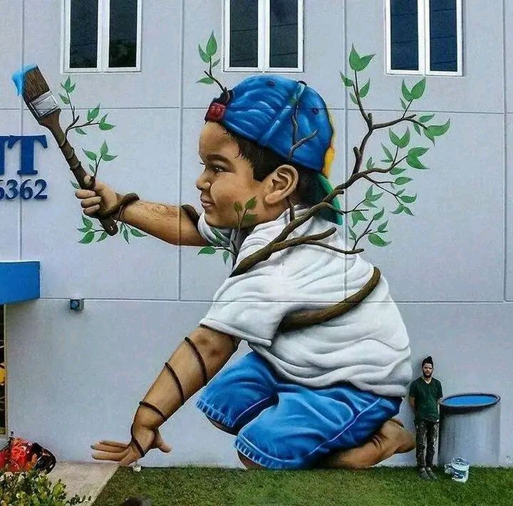 Street art mural of a crouching child in blue painting leafy branches on a white wall