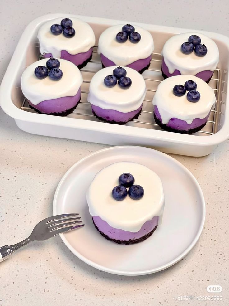 Small purple layered cakes topped with white frosting and fresh blackberries on white tray
