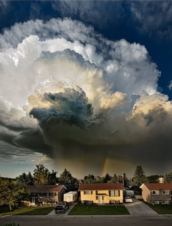 Massive storm clouds above suburban houses with a faint rainbow in the distance