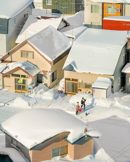 Aerial view of snowy Japanese houses with people shoveling snow from roofs and ground in a residential neighborhood