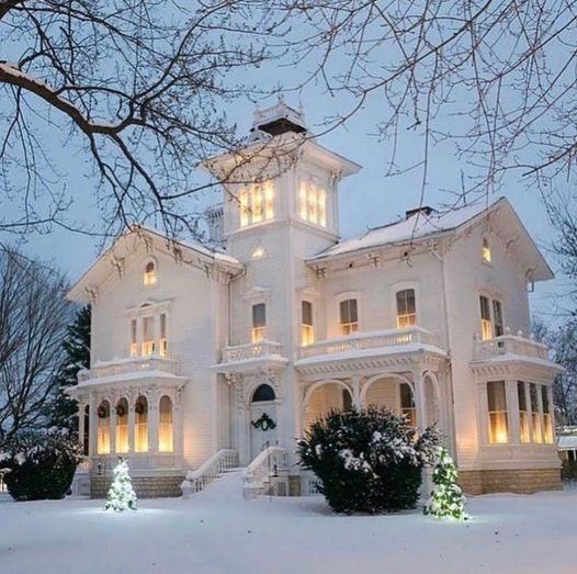 White Victorian house with tower illuminated at twilight in snowy winter landscape with bare trees