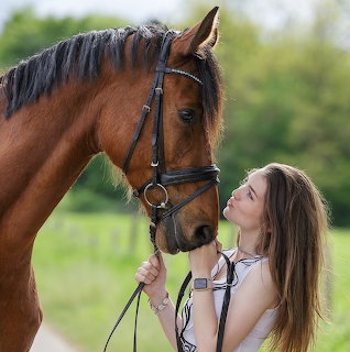 Young woman in white top standing close to brown horse with bridle in green outdoor field