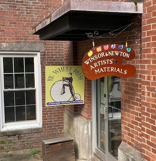 Brick art supply storefront with hanging sign, glass door, and window