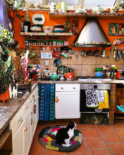 Colorful bohemian kitchen with orange walls, decorated shelves, white appliances, and cat on floor rug