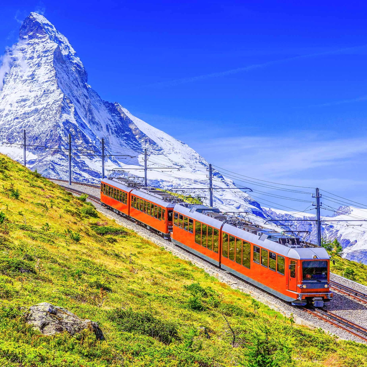 Red train on alpine tracks with snowy Matterhorn mountain peak in background under blue sky