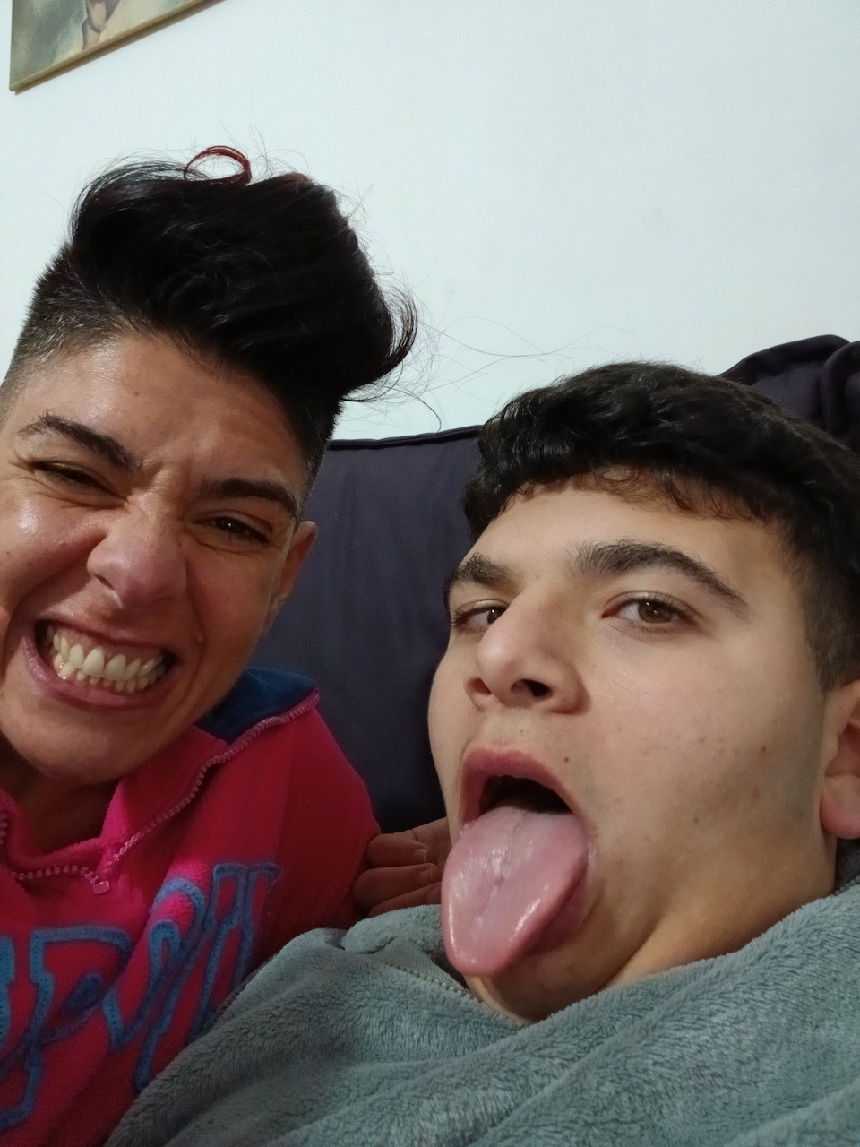 Two young people making funny faces, one sticking tongue out, indoor selfie