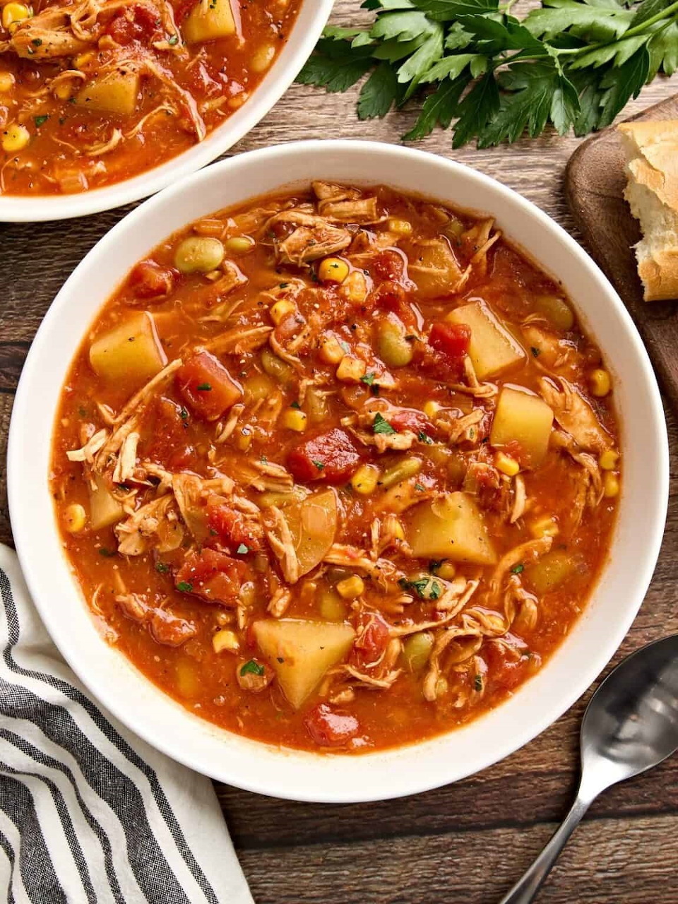 Bowl of chicken Brunswick stew with corn, beans, and tomatoes on wooden surface with parsley garnish