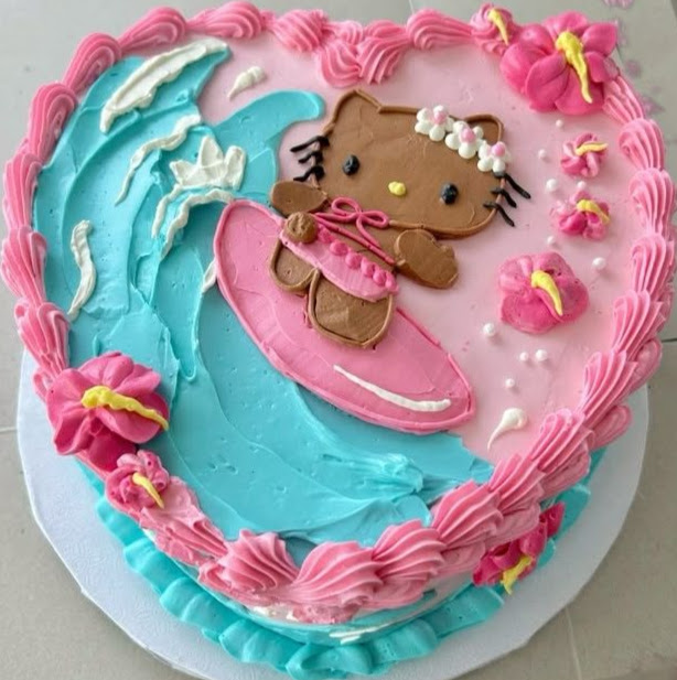 Round cake decorated with Hello Kitty surfing figure, blue wave frosting, pink border, and tropical flower decorations