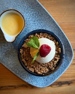Overhead view of a fruit crumble topped with ice cream, raspberry, and mint, accompanied by a small jug of custard