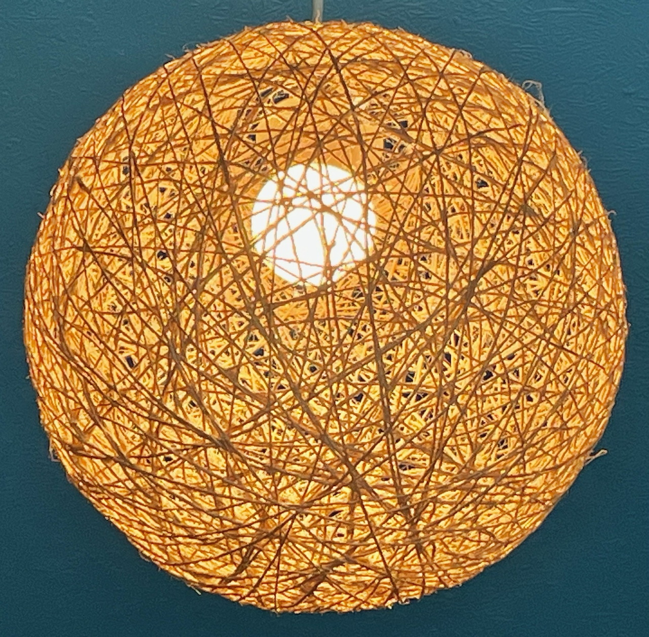 Round woven string lamp glowing warm yellow against teal wall