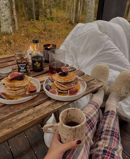 Rustic outdoor breakfast table with pancakes, berries, syrup, coffee, and a cozy blanket-covered chair