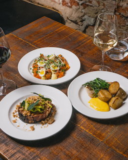 Three white plates with gourmet food on a wooden table with wine glasses and brick wall background