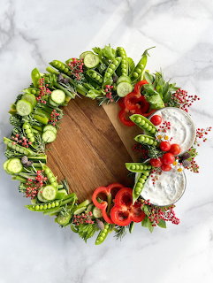 Circular wooden cutting board surrounded by fresh sliced vegetables arranged in wreath shape on white marble