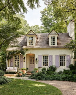 Quaint cottage with lavender shutters, dormer windows, and green shrubs along a garden path