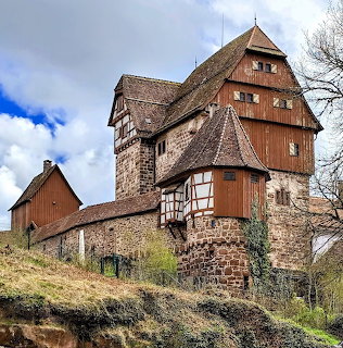 Medieval half-timbered castle with stone walls and tower on hillside under partly cloudy sky
