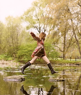 Woman in patterned dress and boots jumping across stones over pond water with trees in background