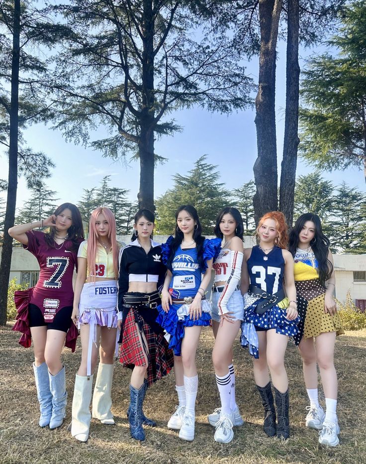 Seven K-pop girl group members in colorful outfits posing together outdoors near trees