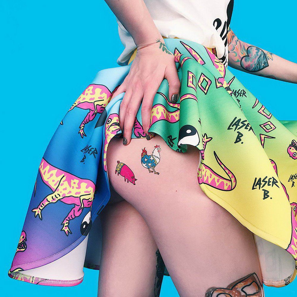 Woman in colorful printed skirt with cartoon animal designs showing tatooed leg against blue background