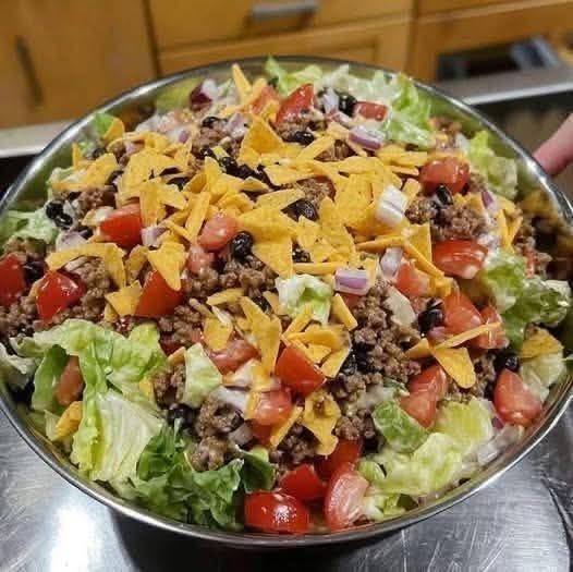 Large metal bowl of taco salad with ground beef, lettuce, tomatoes, black beans, onions, cheese, and Doritos chips
