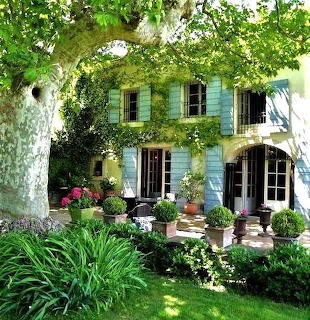 Ivy-covered house with green shutters, arched windows, surrounded by garden with flowers and plants under large tree