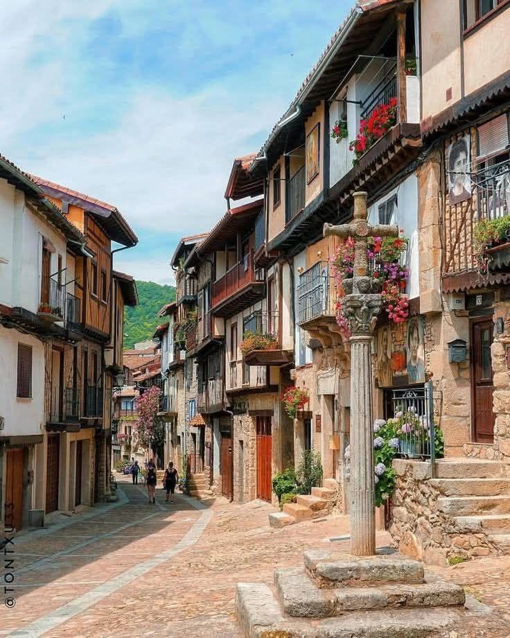 Narrow cobblestone street with old village houses, balconies, and a stone monument