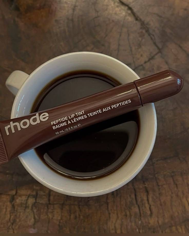 White coffee cup with black coffee and brown Rhode lip gloss on wooden surface
