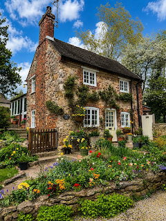 Traditional English stone cottage with blooming garden beds full of spring flowers and climbing vines on sunny day
