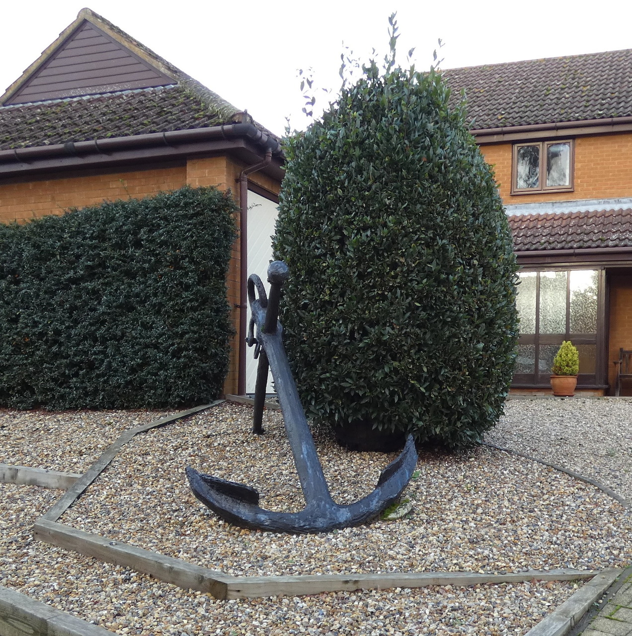 Large anchor sculpture on gravel beside a hedge and brick building