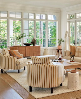 Bright sunroom with striped armchairs, wooden furniture, and floor-to-ceiling windows overlooking lush greenery