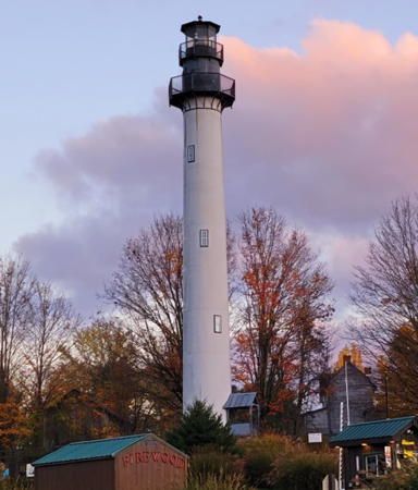 Tall white lighthouse above trees and small buildings under pink clouds
