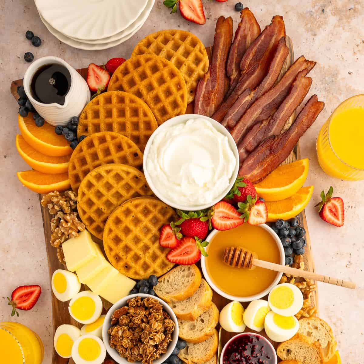 Breakfast charcuterie board with waffles, bacon, boiled eggs, berries, oranges, and honey on a platter
