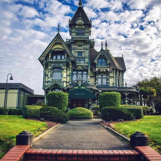 Victorian-style house with ornate rooflines, hedges, and a walkway under a cloudy sky