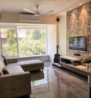Modern living room with grey sectional sofa, stone accent wall, TV, guitar, and large windows with garden view