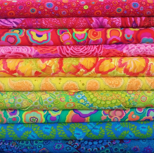 Stack of colorful patterned fabric bolts arranged by rainbow hues from pink to blue with floral and abstract designs