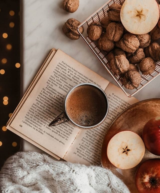 Top-down view of a coffee mug on an open book with walnuts, sliced apples, and a knit blanket on marble
