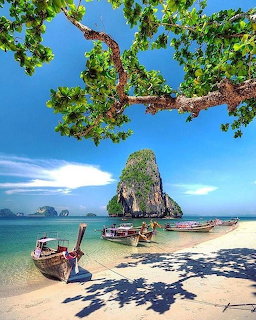Tropical beach with longtail boats, limestone karst island in turquoise sea, tree branches overhead, clear blue sky