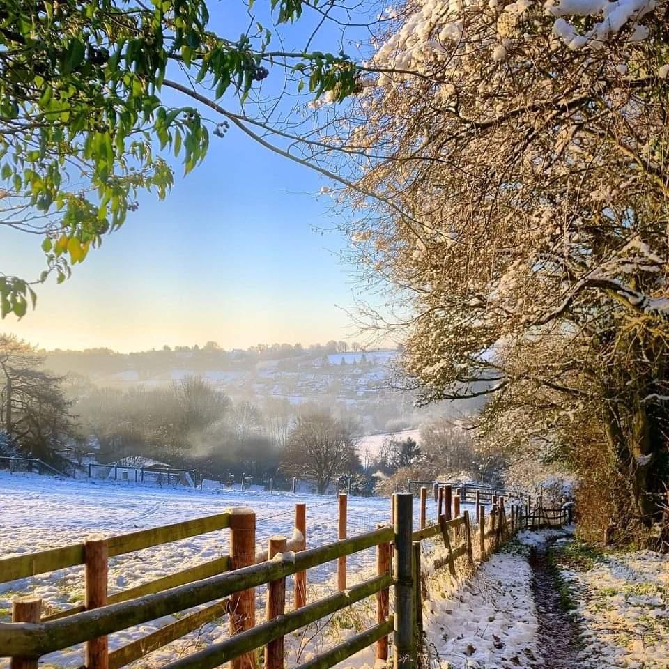 Snowy countryside path with wooden fence leading through winter Cotswolds landscape with rolling hills and village
