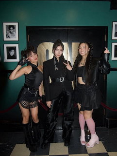 Four women in black outfits posing together in front of a fireplace with teal walls
