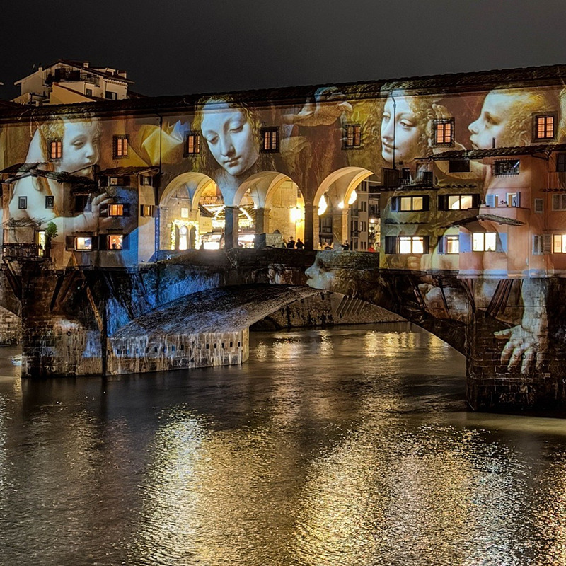 Building facade at night with projected artwork of faces and hands, reflecting in water
