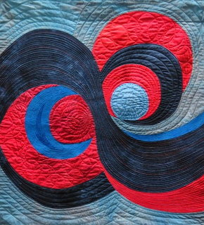 Quilted fabric art with swirling dark navy spirals and red and blue circles on light blue background