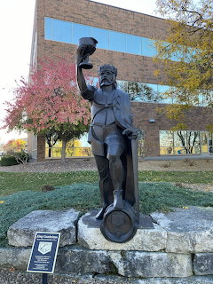 Bronze statue of King Gambrinus raising a goblet, on stone pedestal with autumn tree and building behind