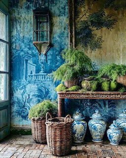 Interior with blue toile painted wall, green plants on rustic table, wicker baskets, and blue white vases