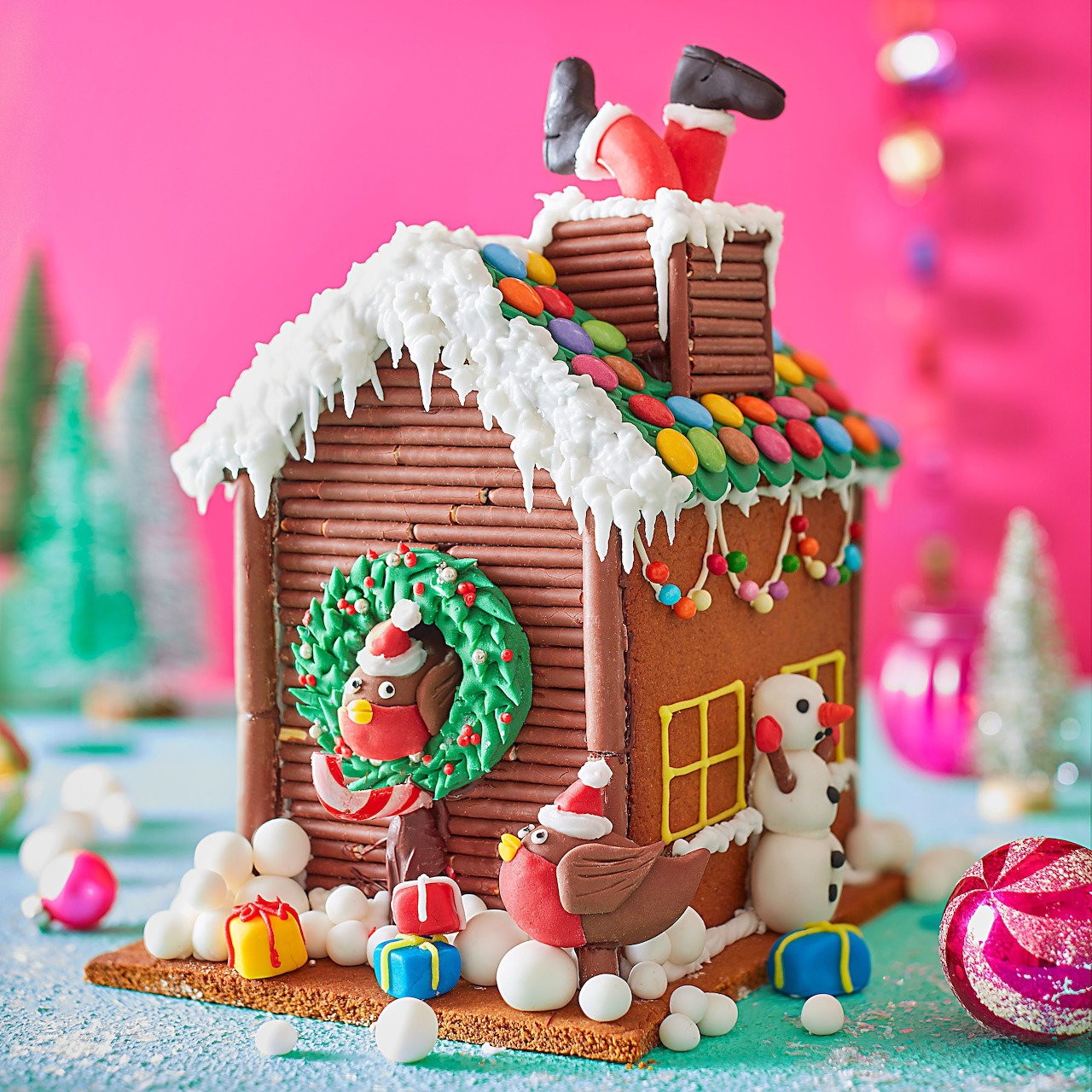 Decorated gingerbread house with icing, candies, wreath and miniature figures against pink background