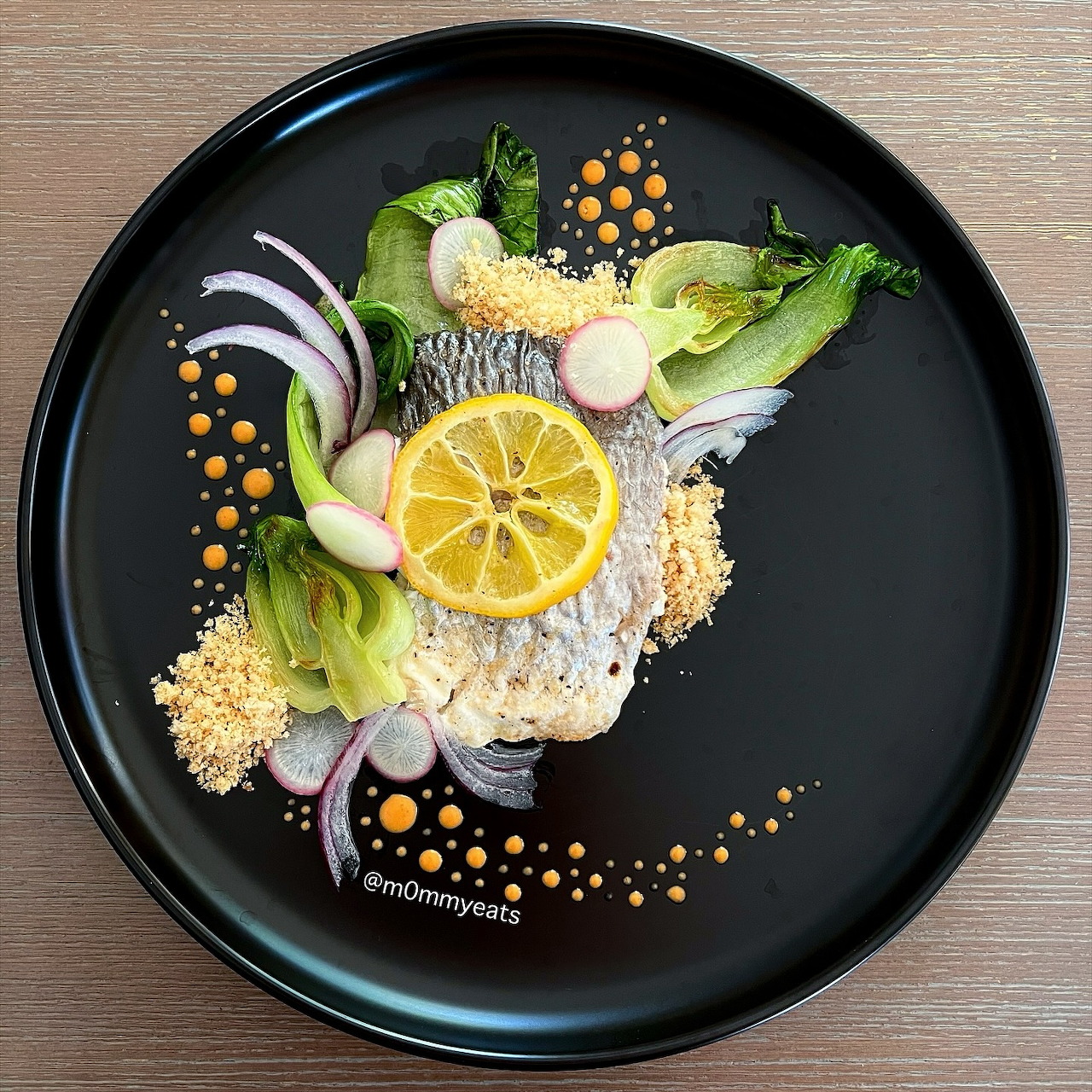 Artfully plated fish with lemon slice, greens, and colorful garnishes on black plate