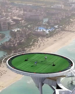 Circular green tennis court on helipad platform high above Dubai coastline with luxury resort and beach below
