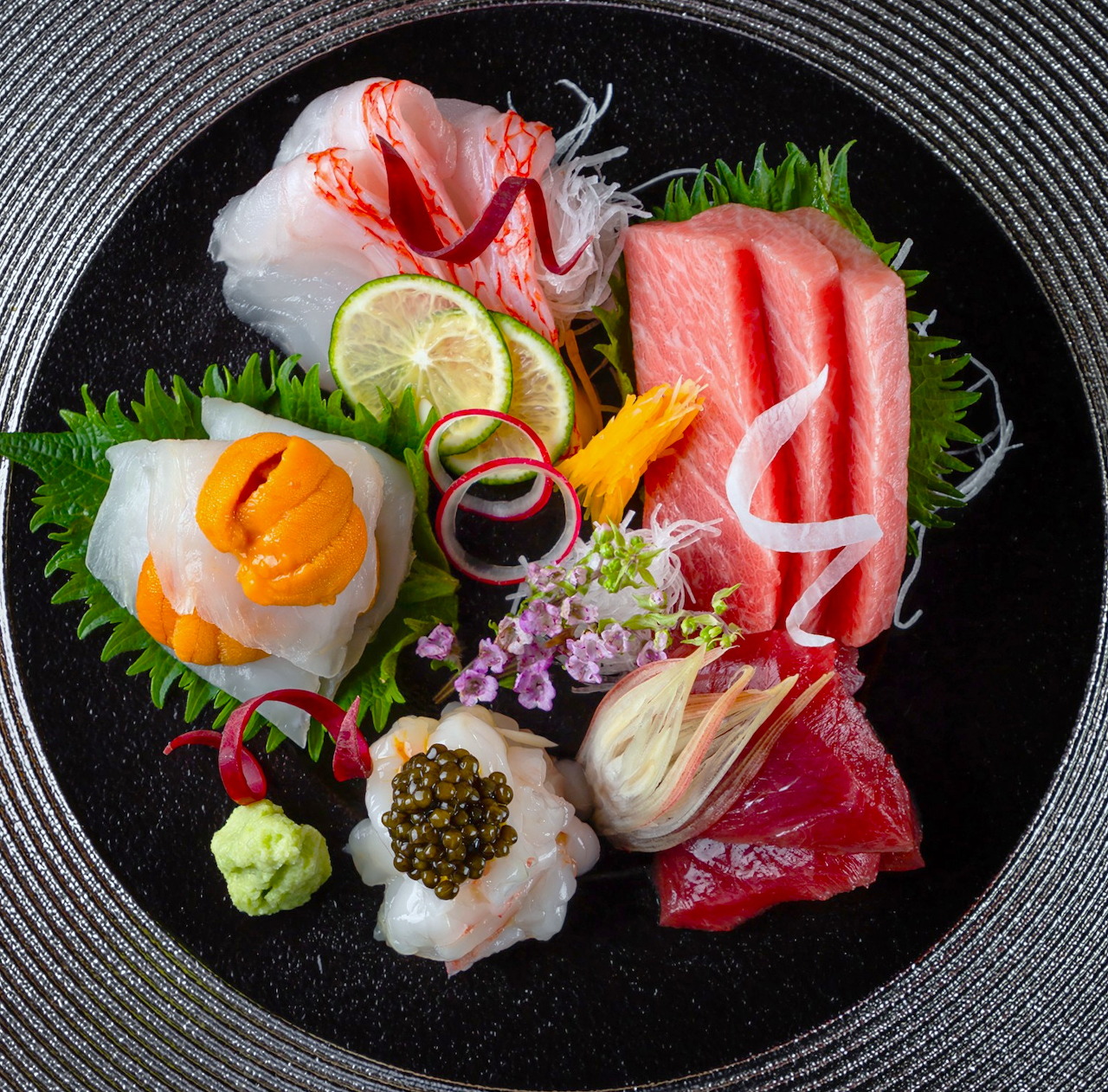 Overhead view of colorful sashimi platter on black plate with fresh raw fish, garnishes, and lemon slices