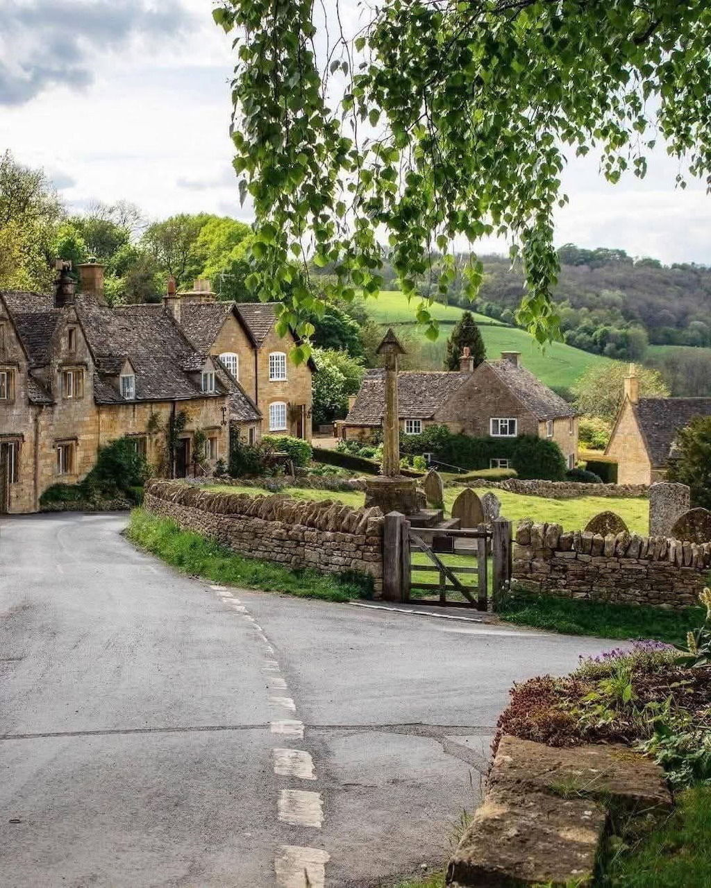 Cotswolds village street with stone cottages, green hills, and a wooden gate along a quiet lane